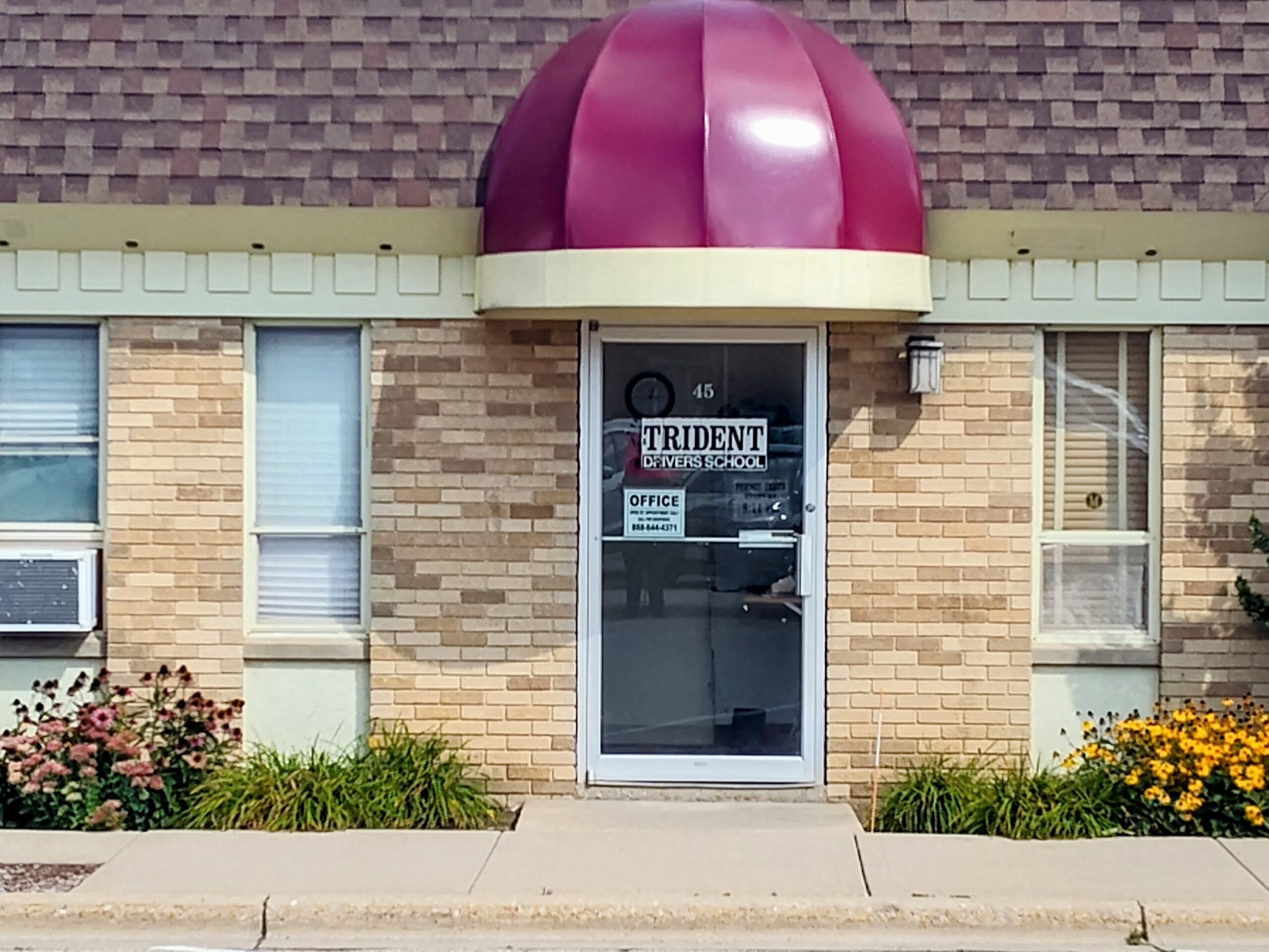 Single-story brick building featuring a maroon awning, large windows, and a "For Rent" sign on the door. Captures local rental property.