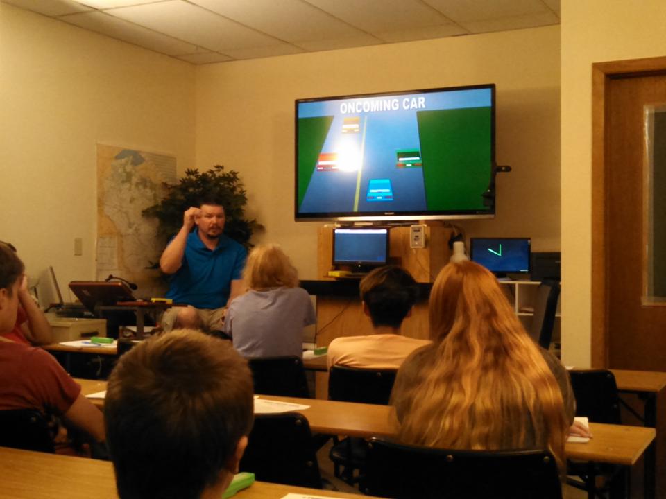 A driving instructor teaches a class about road safety, using a graphic on a screen showing an oncoming car scenario. Students are engaged in learning.