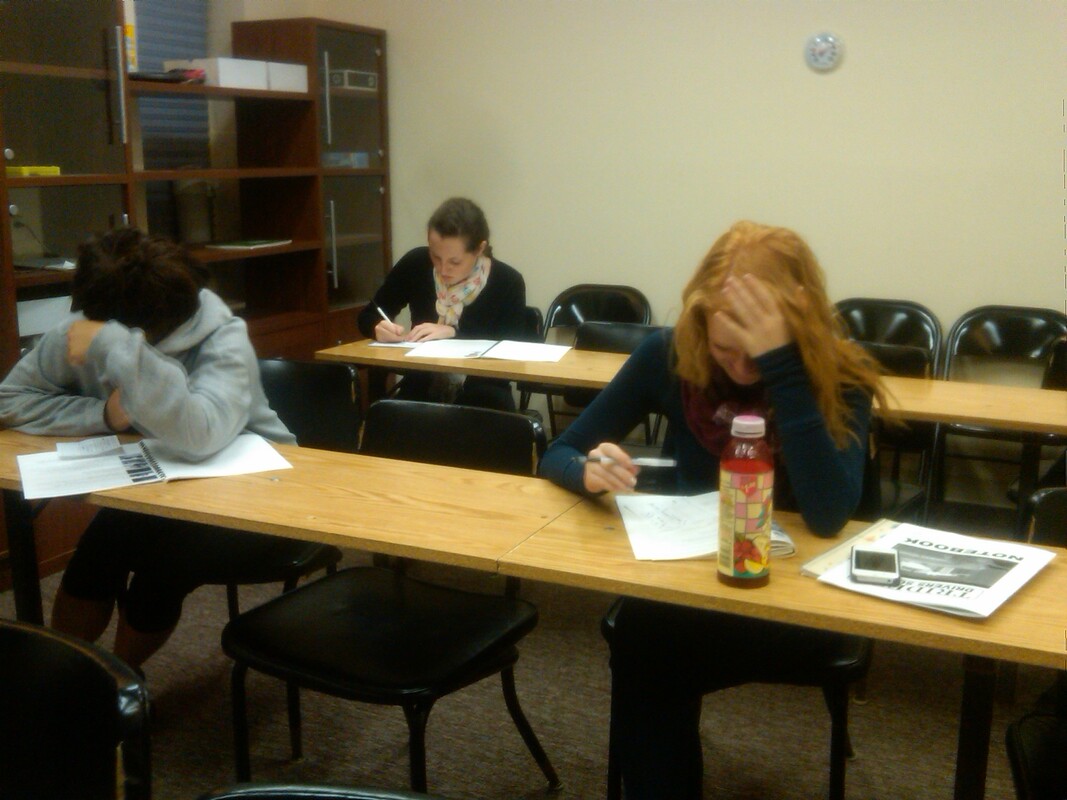 Students in a classroom take an exam, displaying varying expressions of focus and anxiety. One student rests her head on the table, while another writes intently.